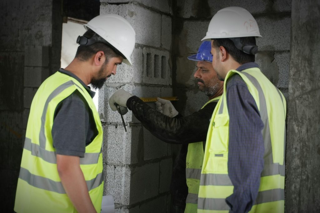 Three construction workers in hi-vis vests and safety helmets measuring a blockwork wall on site